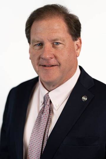Headshot photo of Mark Farley, a middle-aged Caucasian man with brown hair in a short haircut wearing a black suit jacket, white button down, and red patterned tie, with an Upper Cumberland pin on his jacket.